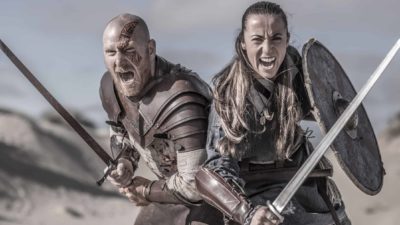 two medieval style warriors wearing armour and carrying shields and swords stand side by side as if joined at the hip with fericious, wide mouthed expressions on their faces as if ready for battle.