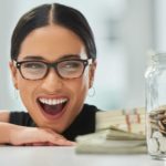A woman wearing glasses and a black top smiles broadly as she stares at a money yarn full of coins representing the rising JB Hi-Fi share price and rising dividends over the past five years