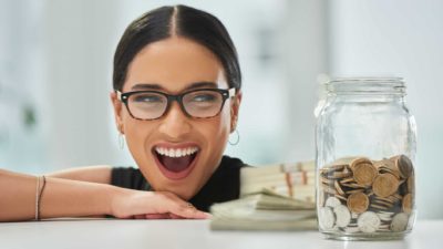 A woman wearing glasses and a black top smiles broadly as she stares at a money yarn full of coins representing the rising JB Hi-Fi share price and rising dividends over the past five years