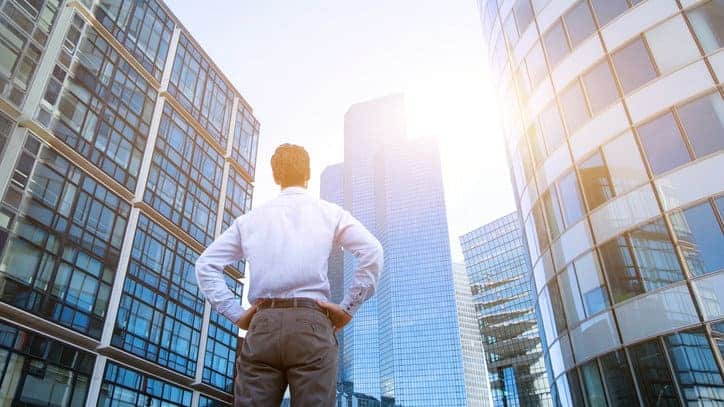 a man dressed in businesswear stands with his back to the camera and hands on hips looking up at high rise buildings in a dense urban setting.