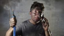 A man witgh a dirty face as though it has been blackened by smoke or a small explosion holds up an electrical wire as he talks on the phone with a worried expression on his face.