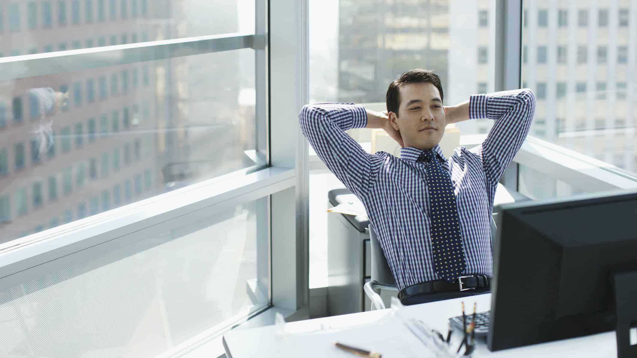 A businessman wearing a shirt and tie leans back in his chair with his hands behind his head while looking at a computer screen in an office with high rise buildings visible out the windows.
