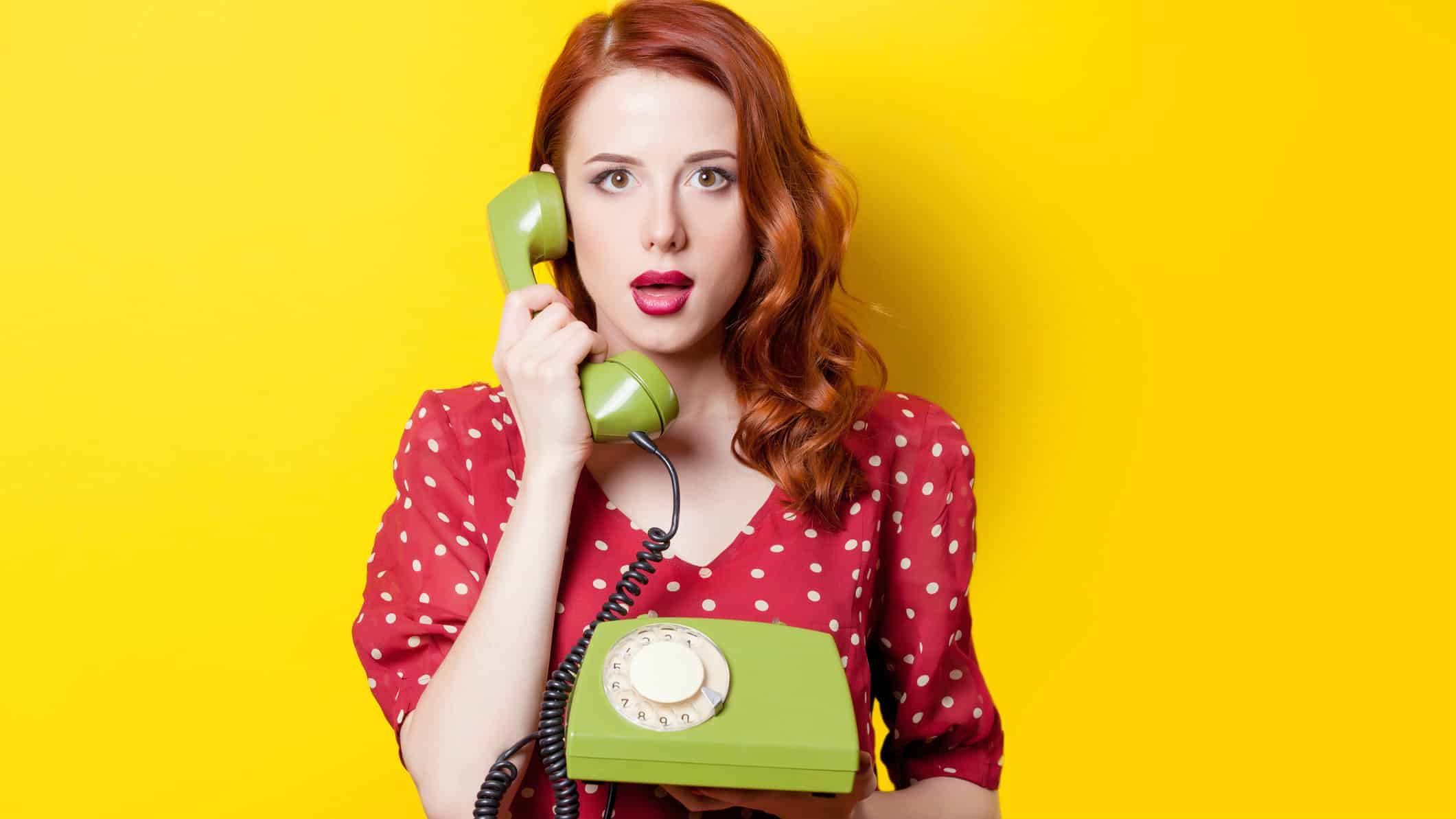 A woman with red hair wearing a red polka-dot dress holding a retro green telephone with the handset at her ear