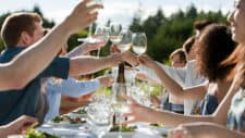 People hold up glasses to cheers across a long dining table outdoors as they sit down to feast.