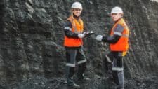 Two miners stand in front of a large black wall of coal.