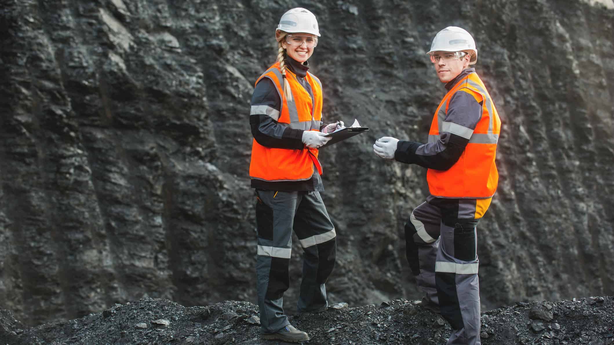 Two miners stand in front of a large black wall of coal.