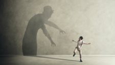 A man runs away from a large shadow on the wall reaching down with its arms as if to grab him.