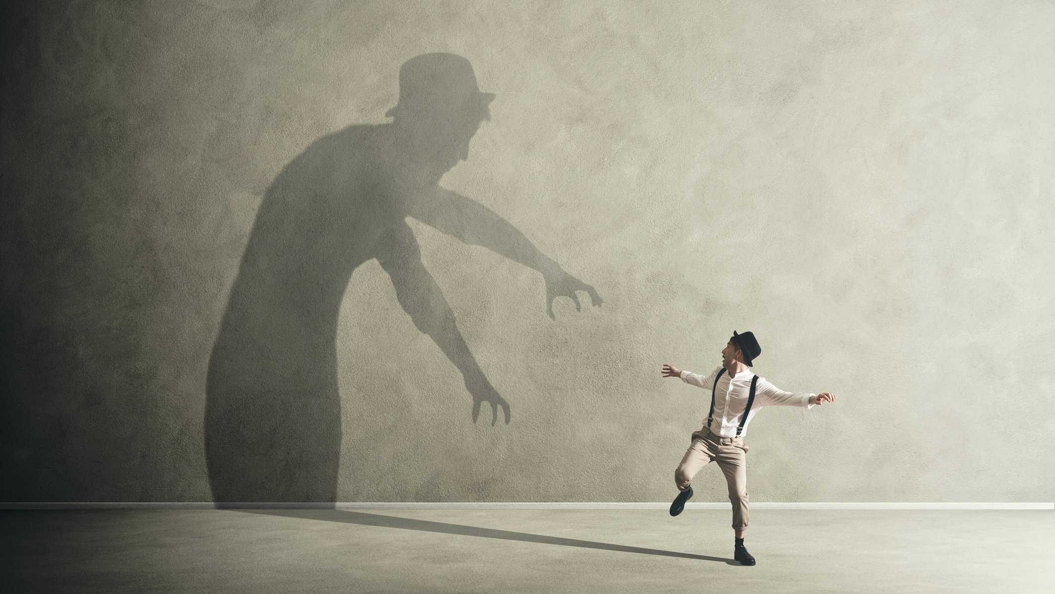 A man runs away from a large shadow on the wall reaching down with its arms as if to grab him.