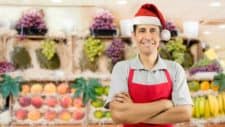A supermarket worker stands in front of a display of fresh produce wearing a red santa hat and apron, smiling widely with his arms folded.