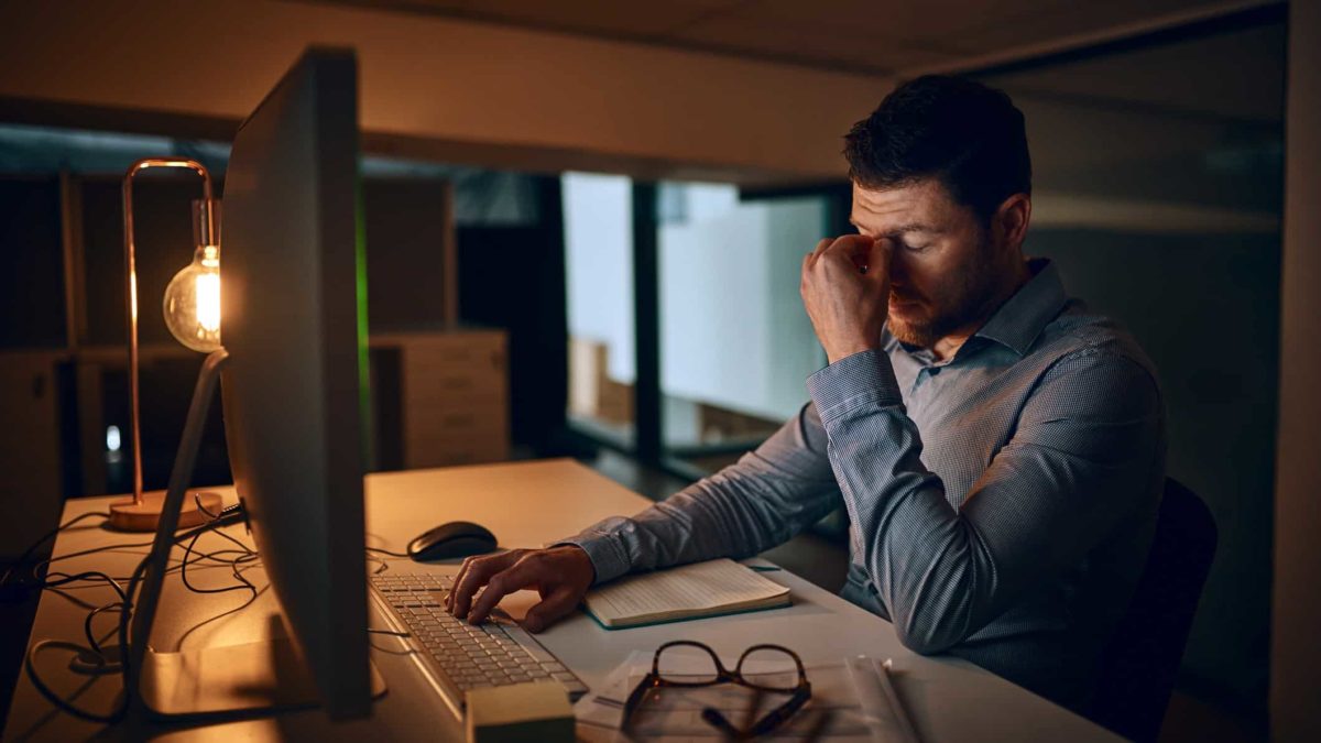 Man struggles to work in dark room at computer, puts head in hand