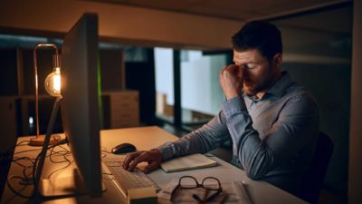 Man struggles to work in dark room at computer, puts head in hand