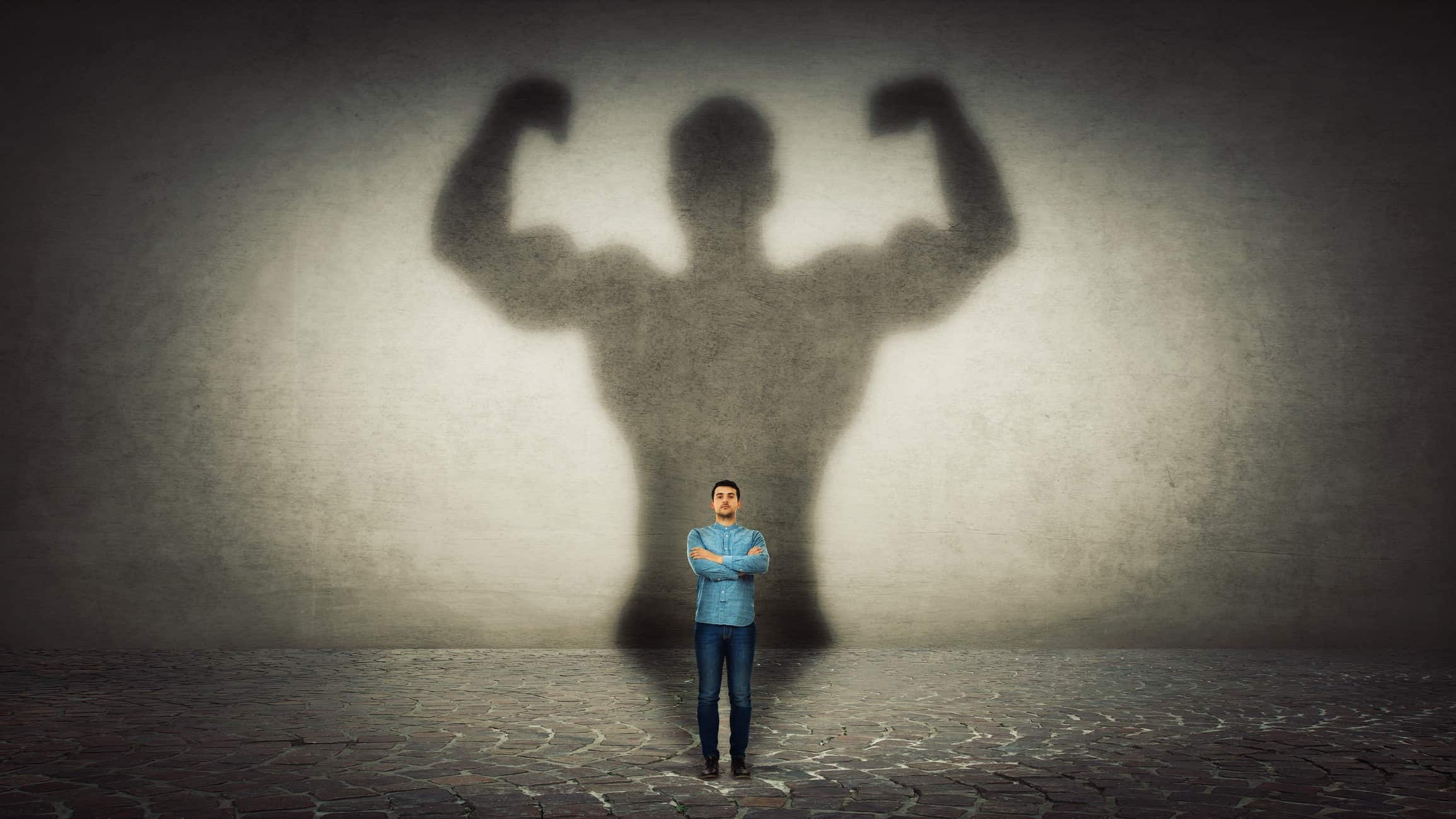 A small man stands with arms crossed confidently as a huge shadow bearing giant biceps looms large behind.