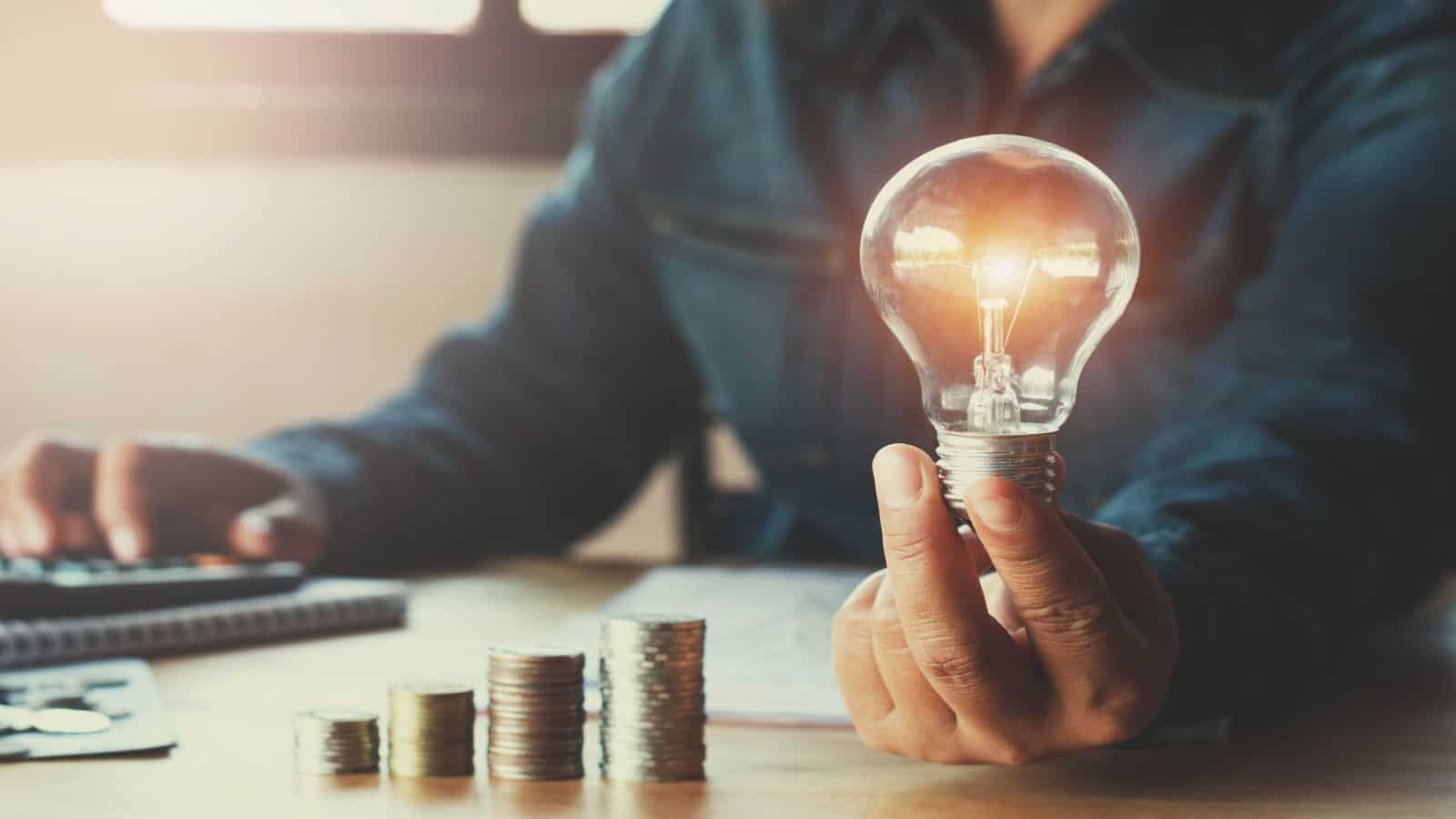 man holding light bulb next to growing piles of coins