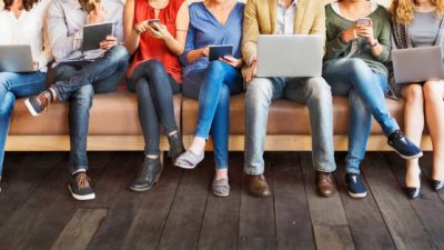 A row of 6 ASX investors sitting on a couch trading ASX 200 shares using various devices phones and laptops trading shares