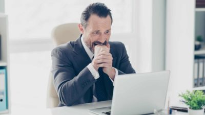 A worried man in a business suit sits at his desk with his fist in his mouth while looking at his laptop