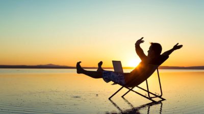 Man in deck chair on a beach at sunset with laptop and arms outstretched