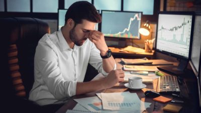 businessman sitting at desk with head in hands in front of computer screens with falling financial charts, asx recession