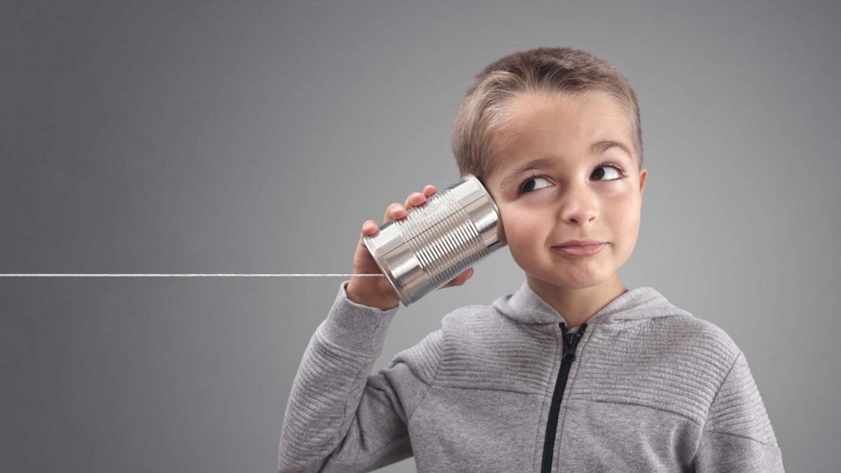A young boy in a grey zip-up jumper has a tin can connected to a string pressed to his ear.