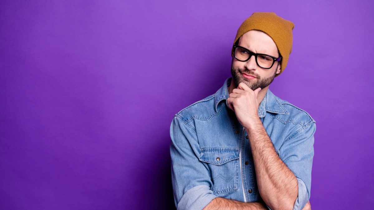 A male ASX investor wearing glasses and a beanie and denim shirt puts his hand to his chin wondering whether to buy ASX shares