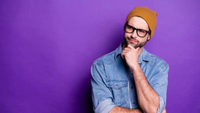A male ASX investor wearing glasses and a beanie and denim shirt puts his hand to his chin wondering whether to buy ASX shares