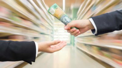 businessman handing $100 note to another in supermarket aisle representing woolworths share price