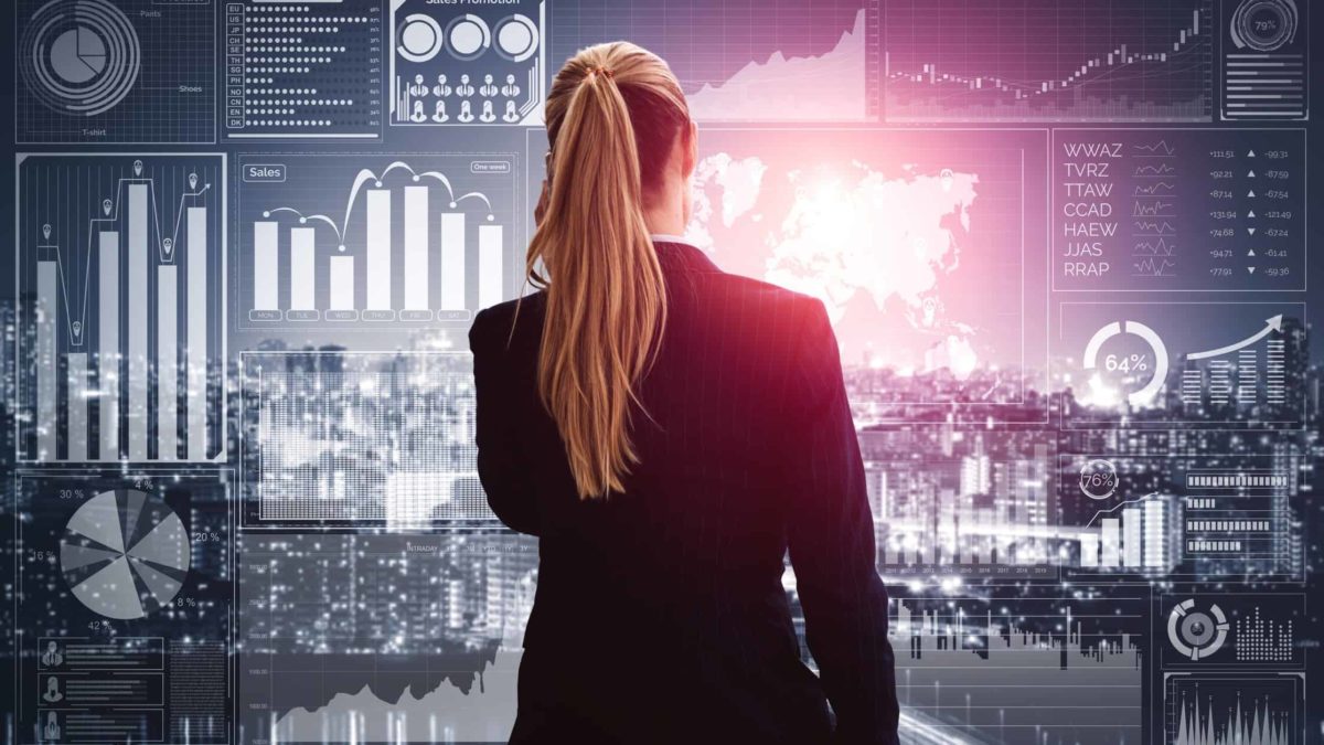 Female ASX investor standing with back to camera, reviewing screen of share price charts in front of her