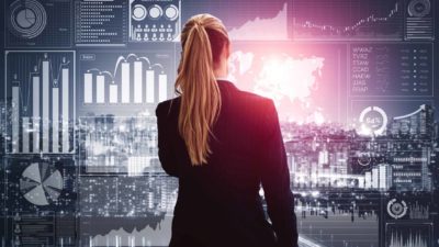 Female ASX investor standing with back to camera, reviewing screen of share price charts in front of her