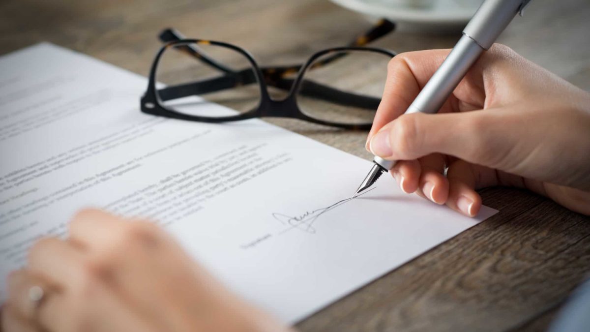 View of hand holding pen signing new deal with glasses sitting on table next to contract papers