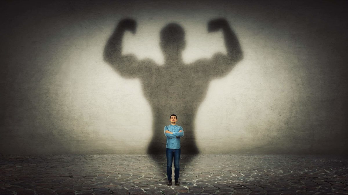 A man stands with arms crossed in front of a giant shadow of a body builder representing ASX small-cap stocks.