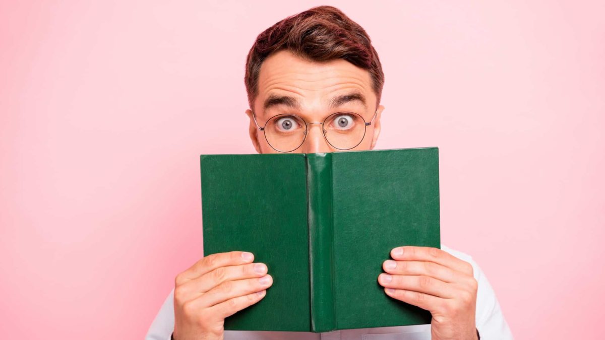 Young male with glasses holding book in front of his face with a surprised expression, indicating a share price movement.