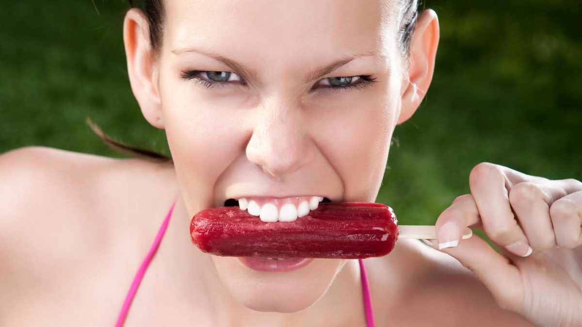 a woman with white teeth sinks them into a frozen ice block and gives a pained expression.