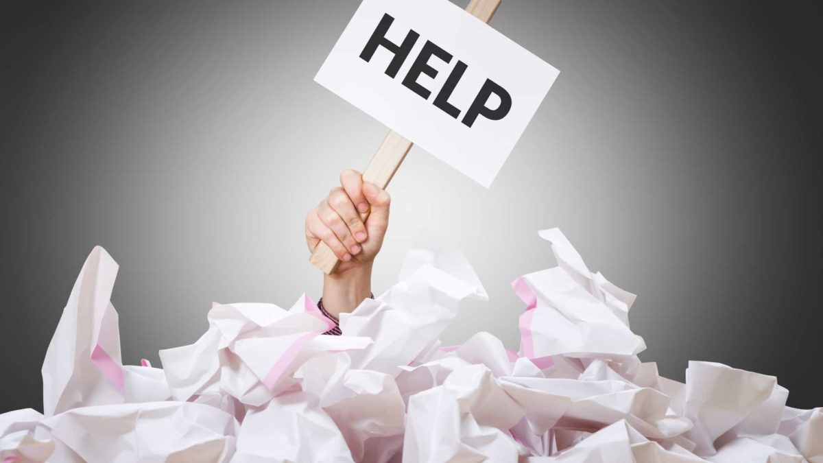 A hand holding a sign which says HELP is buried in paperwork, indicating an overload of information