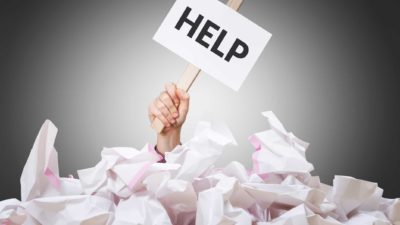 A hand holding a sign which says HELP is buried in paperwork, indicating an overload of information