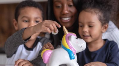 Two kids with their mum put money into a unicorn money box.