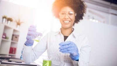 A medical researcher in a white coat holds laboratory equipment and smiles.