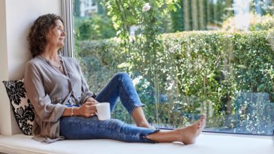 A mature woman looks out her window while drinking a coffee and thinking