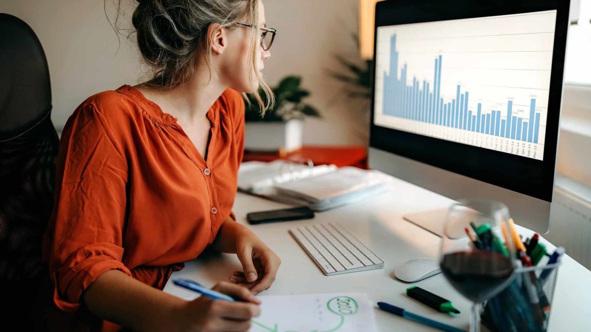 A woman studying share market stats on a computer while writing a report.