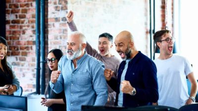 Group of six people in a modern office cheering at a computer screen.