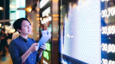 woman with device standing next to large screen displaying rising share price information