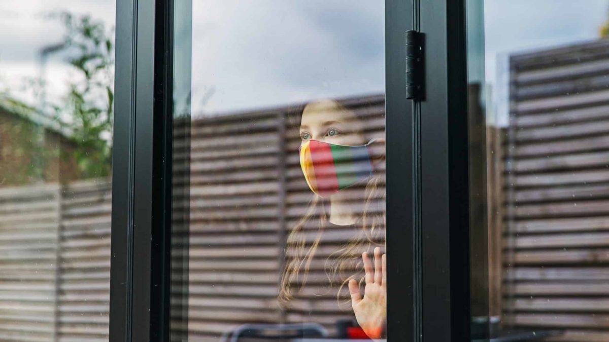 A person wearing face mask looking out a window from indoors.