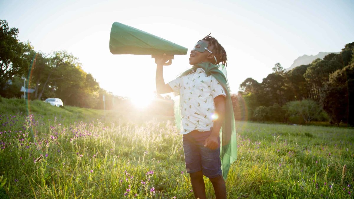 Young boy of African American heritage standing in a field with a green mask and cape shouting through a cardboard megaphone.