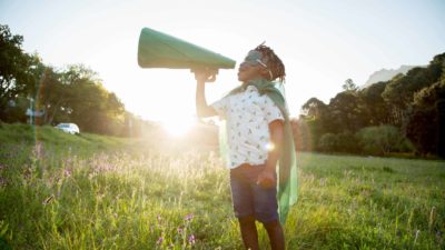 Young boy of African American heritage standing in a field with a green mask and cape shouting through a cardboard megaphone.