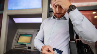 A man looks stressed standing in front of an ATM with his bank card in his hand.