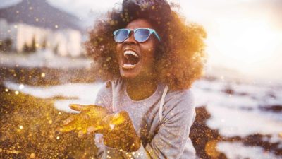 A woman blowing gold glitter out of her hands with a joyous smile on her face.