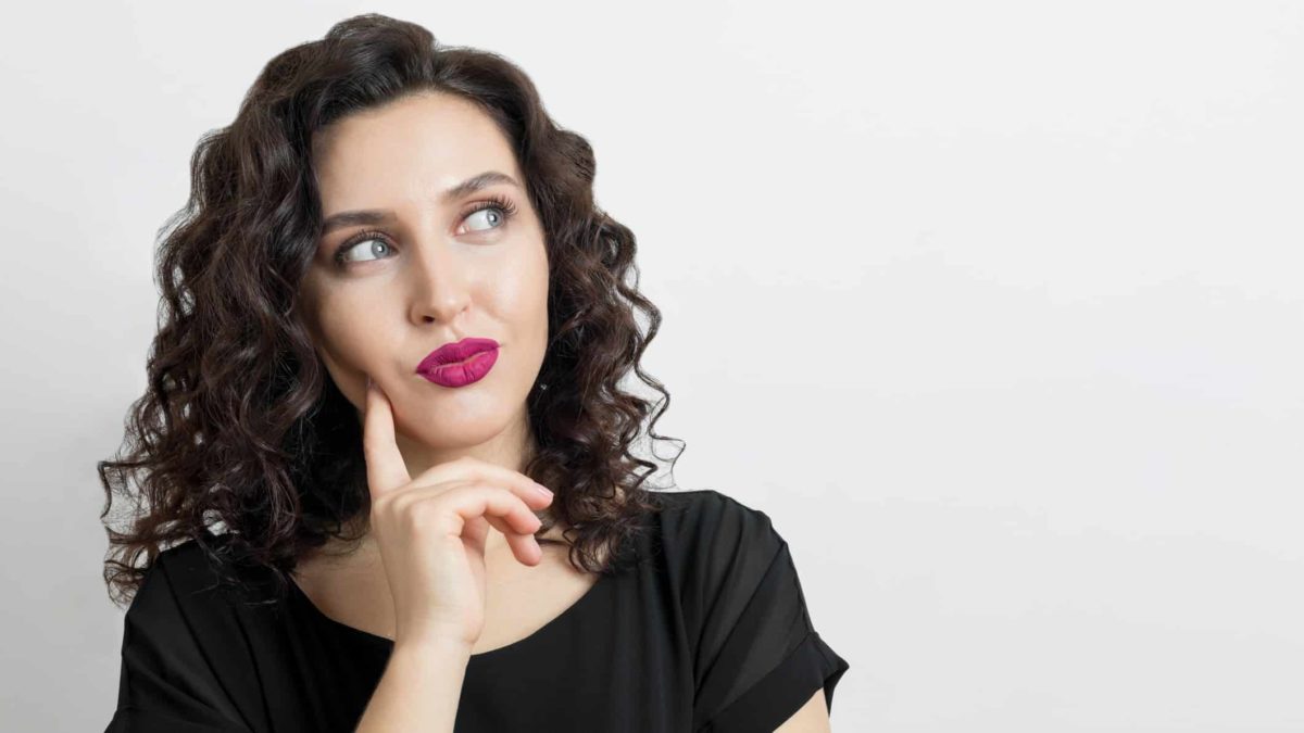 a woman holds her finger to the side of her face and looks upwards as she thinks about something.