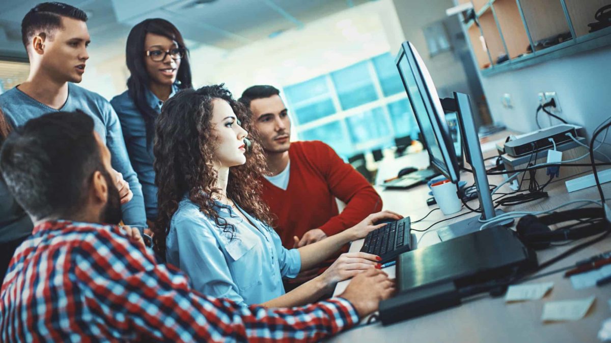 a group of tech people gather around a computer operated by a young woman while the group looks on in support.