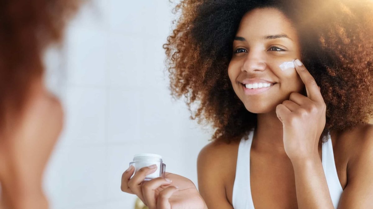 A smiling woman applies face cream to her cheeks while looking in a mirror.