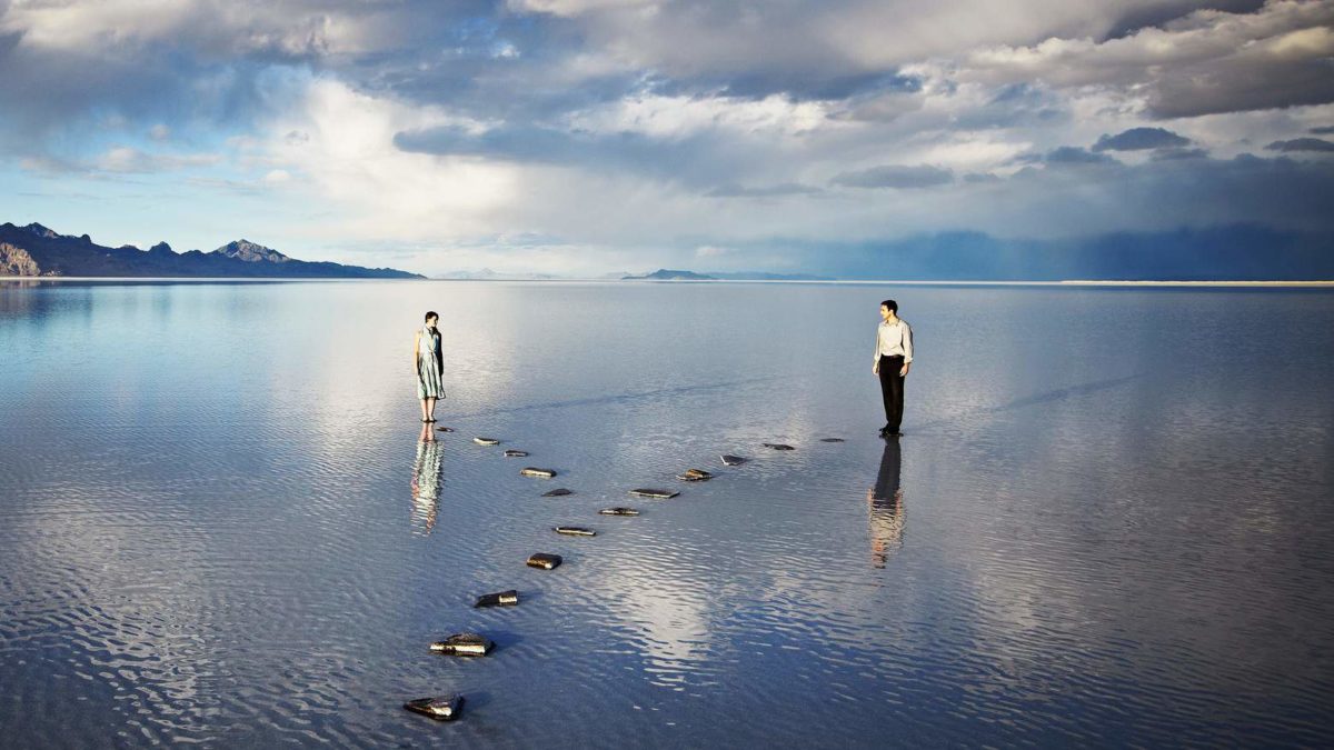 Two people walk in different directions with a vast sky beyond.