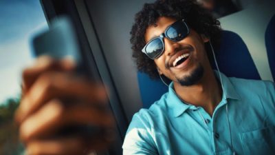 A man in sunglasses is happy with something he's seeing on his mobile phone while sitting on the train.