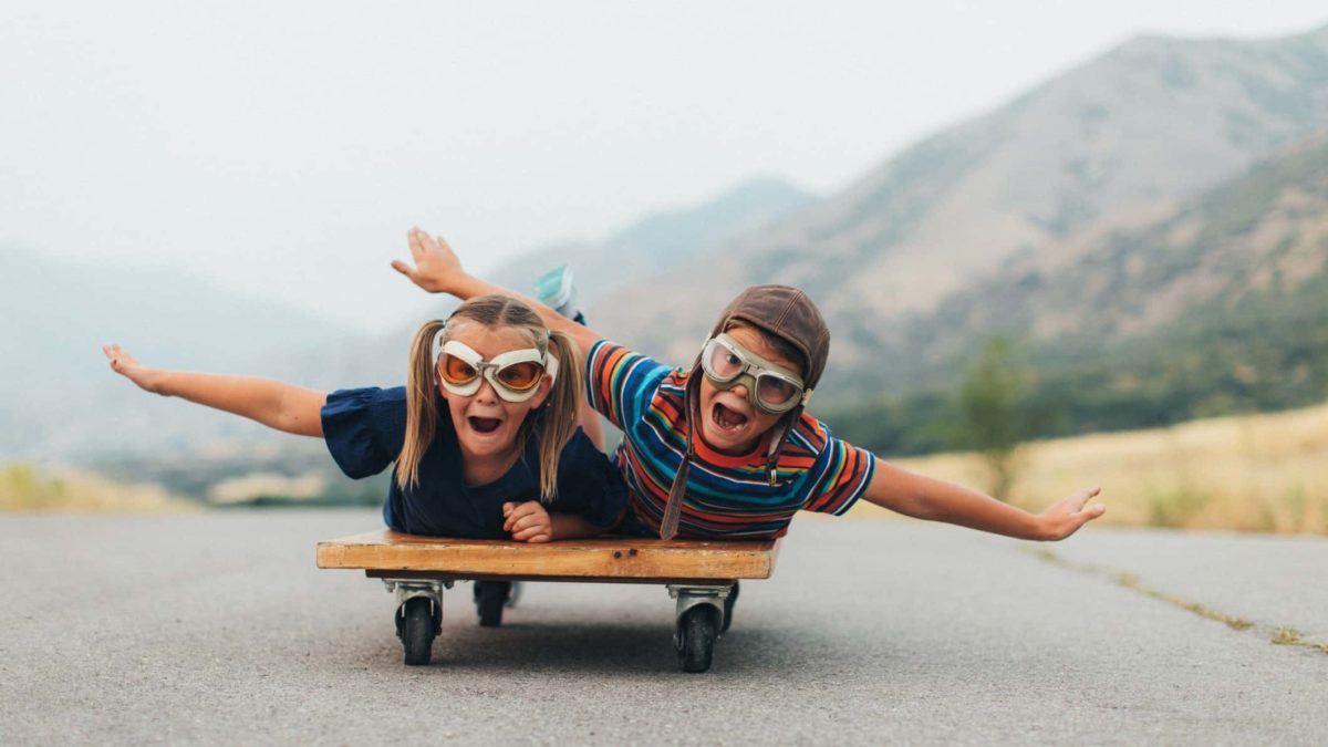 Two kids wearing pilot's goggles take flight down the runway on their tummies with arms outstretched like wings.
